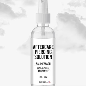 May include: A clear plastic bottle with a white spray nozzle. The label reads "Aftercare Piercing Solution", "Saline Wash", "100% Natural and Gentle", "2FL/6ML", and "Made in U.S.A."