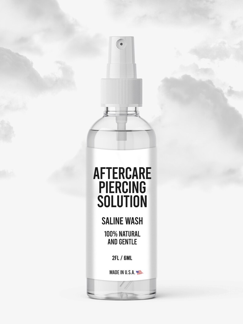 May include: A clear plastic bottle with a white spray nozzle. The label reads "Aftercare Piercing Solution", "Saline Wash", "100% Natural and Gentle", "2FL/6ML", and "Made in U.S.A."