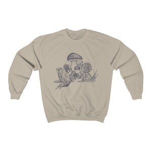 May include: A beige sweatshirt with a blue illustration of a frog, snail, and mushrooms. The illustration is centered on the sweatshirt.