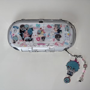 May include: A clear, decorated handheld gaming console with a matching charm. The console features anime-style character art and stickers. The charm has a blue-haired character and pink bead accents. The console is silver and white.