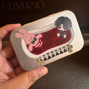 May include: A rectangular silver glitter metal case featuring a printed graphic of a pink rabbit and a dark-haired character sitting in a clawfoot bathtub filled with red liquid. The bathtub has a black and white checkered base and gold feet.