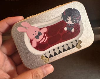 May include: A rectangular silver glitter metal case featuring a printed graphic of a pink rabbit and a dark-haired character sitting in a clawfoot bathtub filled with red liquid. The bathtub has a black and white checkered base and gold feet.