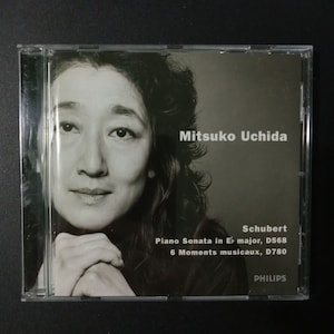 May include: A CD case featuring a black and white photo of Mitsuko Uchida, a Japanese pianist. The text on the case reads "Mitsuko Uchida, Schubert, Piano Sonata in E major, D568, 6 Moments musicaux, D780, PHILIPS".