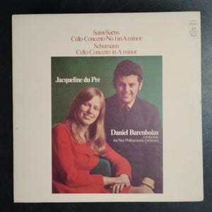 May include: Album cover featuring Jacqueline du Pré and Daniel Barenboim. The cover has a white background with text at the top listing the composers Saint-Saëns and Schumann. The album title is "Cello Concerto". The image shows the two musicians smiling.