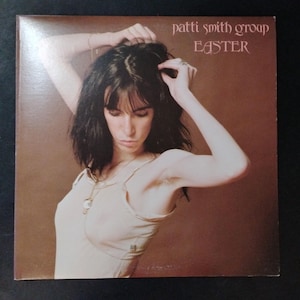 May include: Album cover for Patti Smith Group's "Easter." The cover features a woman with dark hair and a light-colored top, her arms raised. The album title and artist name are in a serif font against a brown background.