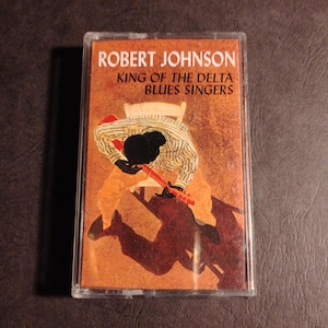 May include: A cassette tape featuring Robert Johnson King of the Delta Blues Singers on the cover. The artwork shows a person in a striped shirt and light pants playing a red guitar while seated on a chair, set against a textured brown background.