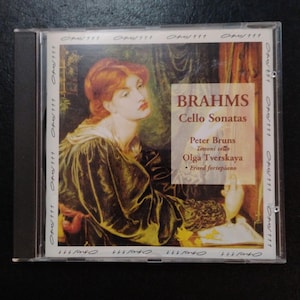 May include: A CD case featuring the album "Brahms Cello Sonatas." The cover art showcases a classical painting of a woman with red hair, wearing a dark gown. The CD case is clear with the text "Opus 111" on the side.
