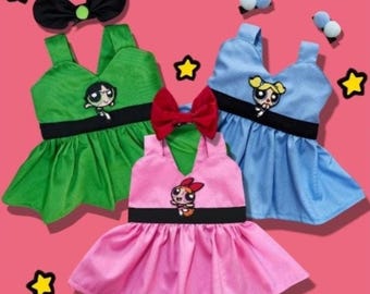 May include: Three sleeveless pet dresses in green, blue, and pink, each featuring a black waistband and a Powerpuff Girls character graphic. The set includes a black bow, a red bow, and two small blue and white pom-pom hair accessories. Text reads The Powerpuff Pets.
