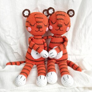 May include: Two crocheted tiger plushies with orange and brown stripes. They have closed eyes and pink cheeks. The tigers are holding hands and have white feet.