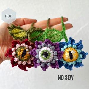 May include: Three crocheted flower ornaments in red, purple, and blue, each with a central eye design. The flowers have detailed petals and are connected to green leaf accents. The text "NO SEW" is visible.