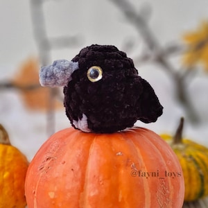May include: A small black crocheted bird with a gray beak sits on top of an orange pumpkin. The pumpkin is on a table with a blurred background of branches and leaves.