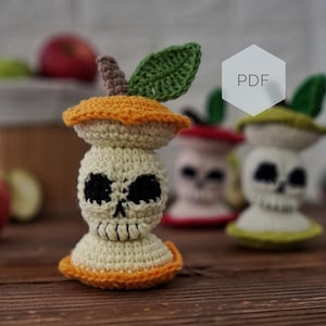 May include: A crochet apple core with a skull face, featuring a cream-colored center, orange top and bottom, and a green leaf. The text PDF is visible in a white hexagon. The item is crafted with yarn and includes black embroidered eye and nose details.