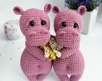 May include: Two crocheted pink hippopotamus plushies holding a bouquet of dried flowers. The hippos have black eyes and are standing on a white surface.