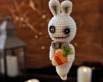 May include: A white crocheted ghost bunny holding a carrot. The bunny has black button eyes and a stitched-on mouth. The carrot is orange and green.