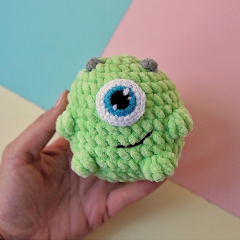 May include: Green crocheted plush toy with a single large blue eye and a small black mouth. The toy is shaped like a monster and has a fuzzy texture.