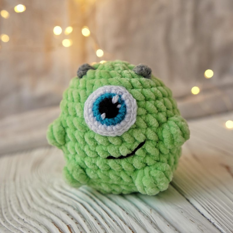 May include: A green crocheted plush toy with a single large blue eye and a smiling mouth. The toy is made with a soft, fuzzy yarn.