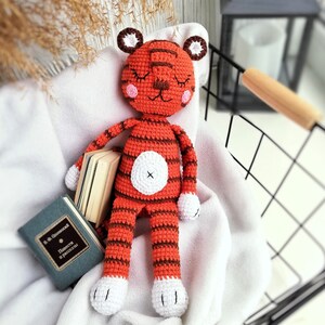 May include: A crocheted tiger stuffed animal with orange and brown stripes, white paws, and a white face with closed eyes. The tiger is lying on a white blanket.