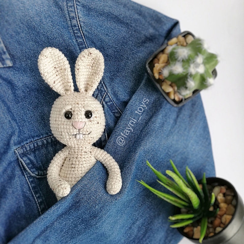 May include: A crocheted cream-colored bunny rabbit with black eyes and a pink nose. The bunny is sitting in a blue denim pocket.