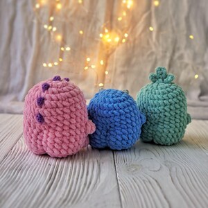 May include: Three crocheted stuffed toys in pink, blue, and green. The toys are sitting on a white wooden surface.