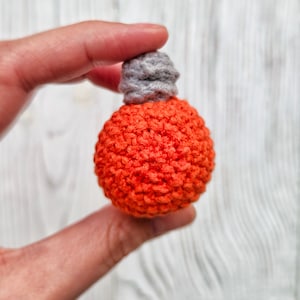 May include: A crocheted orange ornament with a gray crocheted top. The ornament is being held by a hand.
