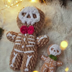May include: Two crocheted gingerbread men, one large and one small, with skull faces and skeleton details. The larger one has a pink bow tie, and the smaller one has a green bow tie. They are brown with white accents, set against a fluffy white background.