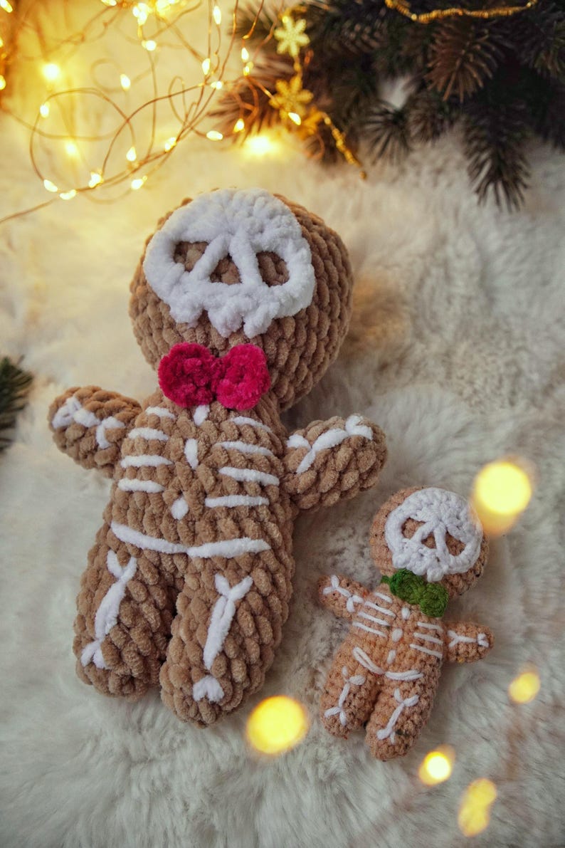May include: Two crocheted gingerbread men, one large and one small, with skull faces and skeleton details. The larger one has a pink bow tie, and the smaller one has a green bow tie. They are brown with white accents, set against a fluffy white background.
