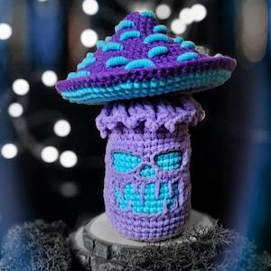 May include: A handmade crochet mushroom in shades of purple and blue. The mushroom cap is a deep purple with blue accents, and the stem features a skull-like face with blue eyes. The mushroom is displayed on a wooden slice.