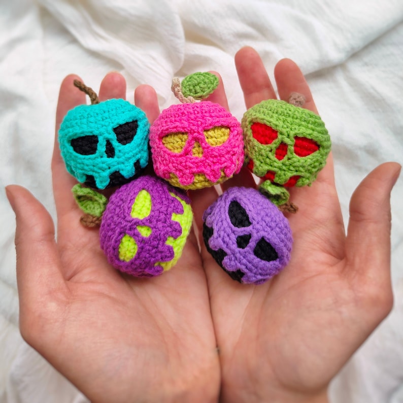 May include: Five crocheted skull-shaped ornaments in different colors, including pink, purple, green, blue, and red. The ornaments are shaped like apples with a stem and leaf on top.