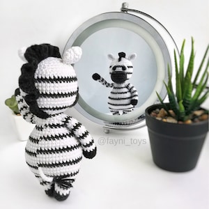 May include: A black and white striped crocheted zebra toy looking at its reflection in a mirror. The zebra has a black mane and tail. The mirror is round and has a silver frame. The text "@fayni_toys" is visible below the mirror.