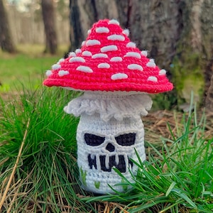 May include: A crocheted mushroom with a red cap dotted with white spots. The mushroom's stem is white with a skull face design and black eyes. Green grass surrounds the mushroom, with a blurred forest background.