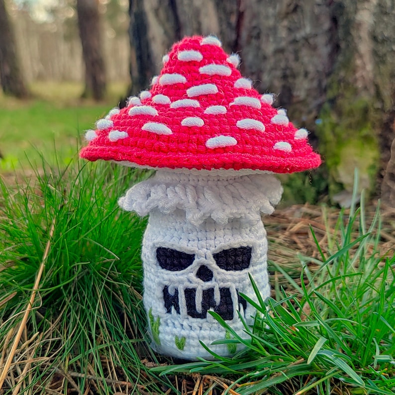 May include: A crocheted mushroom with a red cap dotted with white spots. The mushroom's stem is white with a skull face design and black eyes. Green grass surrounds the mushroom, with a blurred forest background.