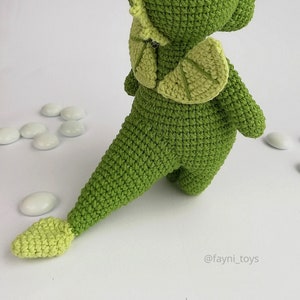 May include: Green crocheted dinosaur toy with a light green tail and wings. The toy is standing on a white surface.