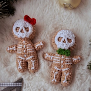 May include: Two crocheted gingerbread skeleton ornaments. One has a red bow, the other a green bow tie. Both have white skull faces and white rib cages. The ornaments are tan and sit on a white, fluffy surface.