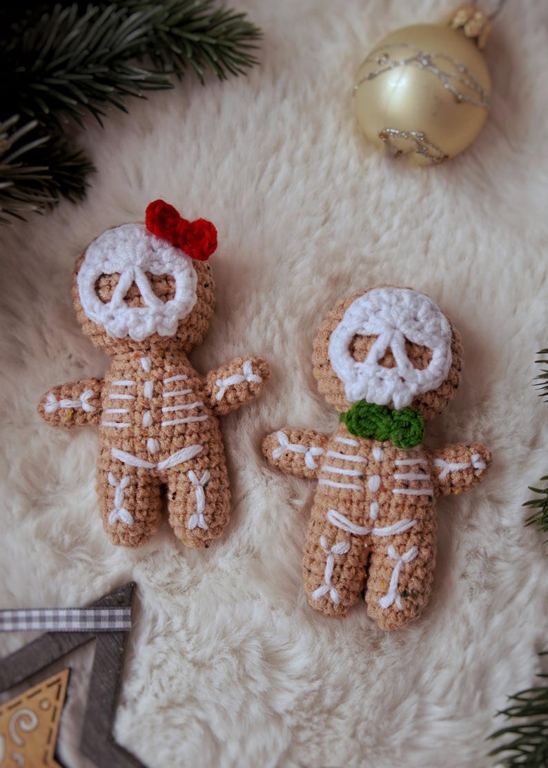 May include: Two crocheted gingerbread skeleton ornaments. One has a red bow, the other a green bow tie. Both have white skull faces and white rib cages. The ornaments are tan and sit on a white, fluffy surface.