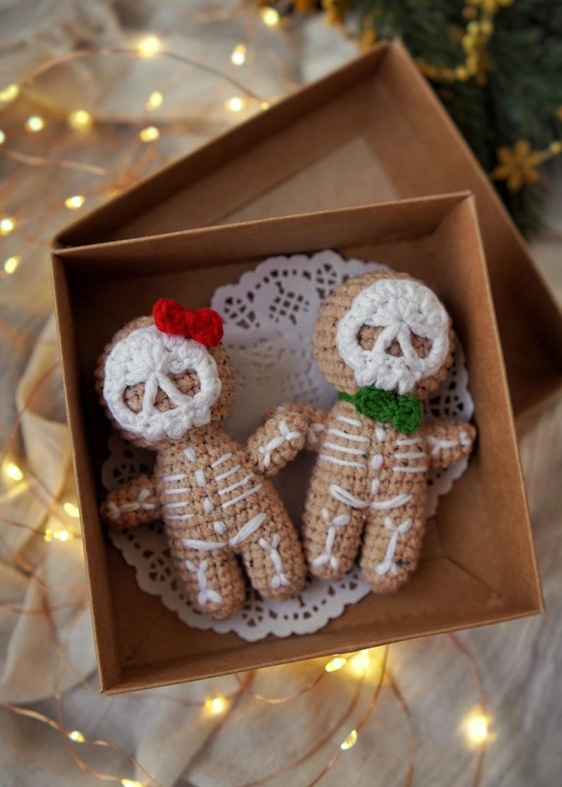 May include: Two crocheted skeleton dolls in a brown box, resting on a white doily. The dolls are beige with white bone detailing and skull faces. One doll has a red bow, the other a green scarf. The box is open, revealing the dolls.