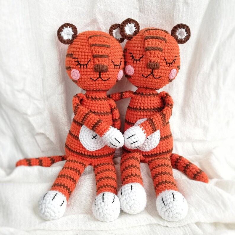 May include: Two crocheted tiger plushies with orange and brown stripes. They have closed eyes and pink cheeks. The tigers are holding hands and have white feet.