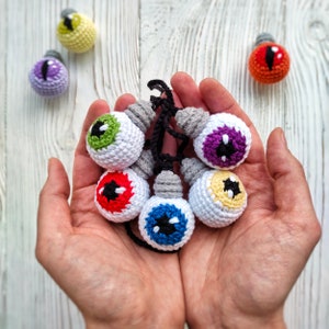 May include: A set of six crocheted eyeball ornaments with different colored pupils, connected by a string. The eyeballs are white with a black outline and are held in a person's hands.