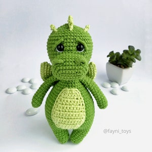 May include: A green crocheted dragon toy with black eyes and a light green belly. The dragon is standing on a white surface with white candy-like objects scattered around it.