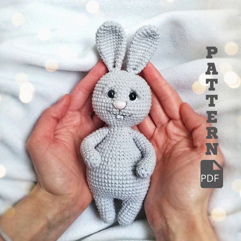 May include: A crocheted gray bunny rabbit toy with black eyes and a pink nose. The bunny is being held in the palms of two hands.