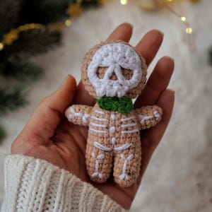 May include: A crocheted skeleton doll with a tan body, white skull face, and green bow tie. The doll's skeleton is outlined in white yarn. The doll is held in a person's hand, with a white sweater sleeve visible in the background.