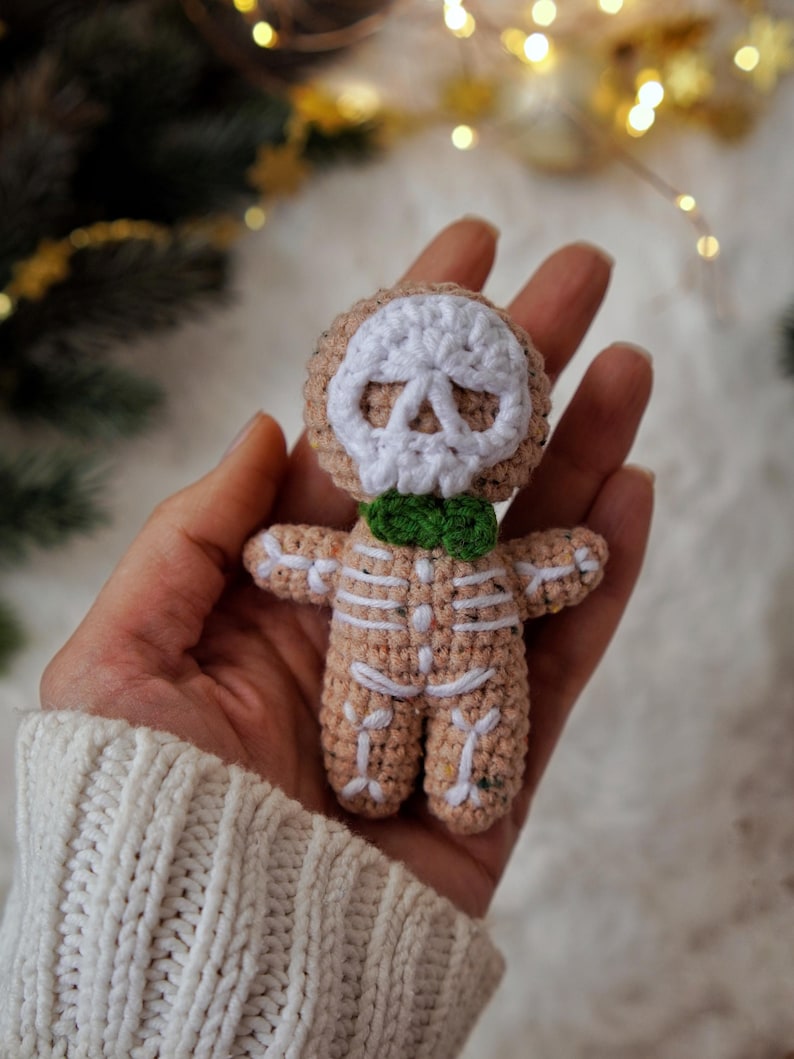 May include: A crocheted skeleton doll with a tan body, white skull face, and green bow tie. The doll's skeleton is outlined in white yarn. The doll is held in a person's hand, with a white sweater sleeve visible in the background.