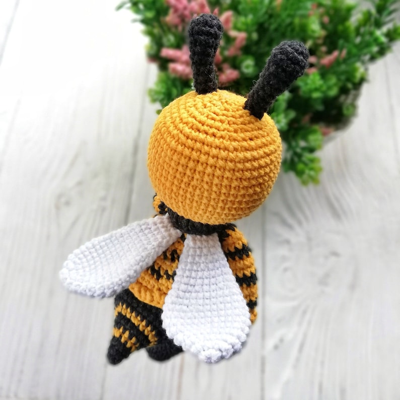 May include: A crocheted bee toy with black and yellow stripes, white wings, and black antennae. The bee is sitting on a white wooden surface.