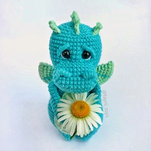 May include: A crocheted blue dragon holding a white daisy. The dragon has green wings and a sad expression. The text '@fayni_toys' is visible in the lower right corner of the image.