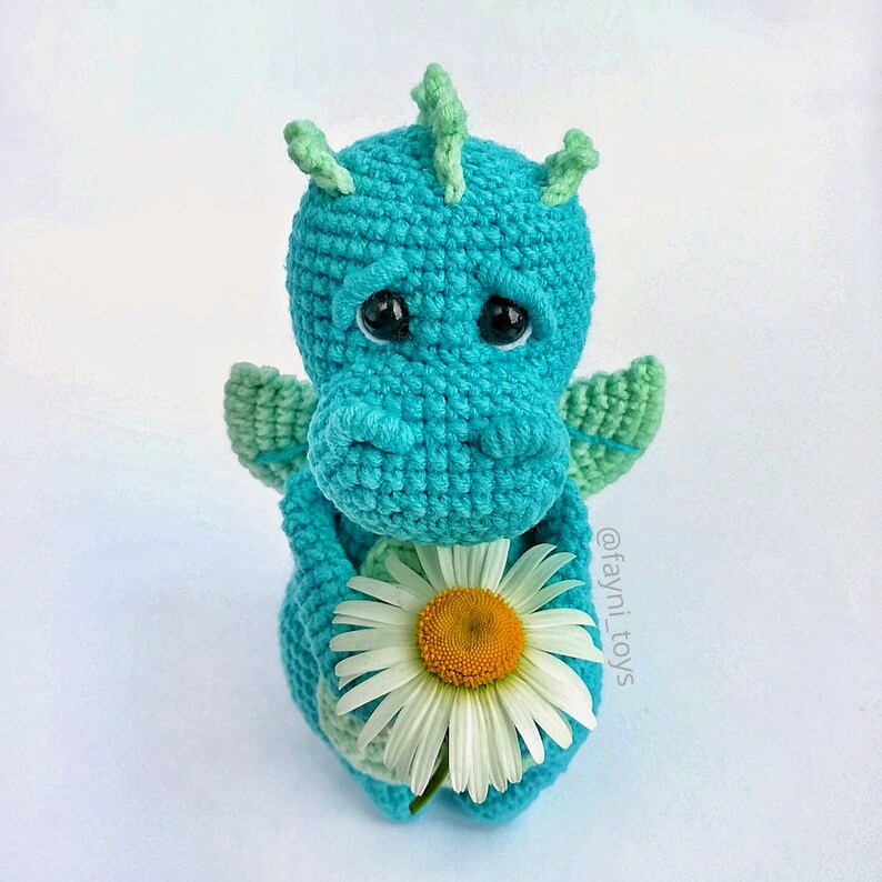 May include: A crocheted blue dragon holding a white daisy. The dragon has green wings and a sad expression. The text '@fayni_toys' is visible in the lower right corner of the image.