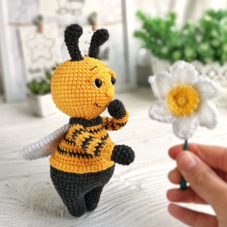 May include: A crocheted bee toy with black and yellow stripes, holding a white crocheted flower. The bee has a black head and a friendly face.