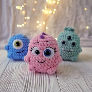 May include: Three crocheted plush toys in the shape of monsters. The first monster is blue with one large eye. The second monster is pink with one large eye and a small purple detail on top of its head. The third monster is green with three eyes.