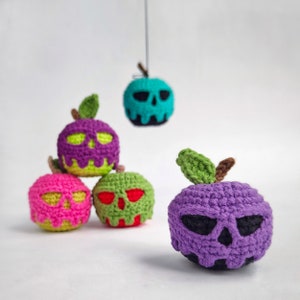 May include: Five crocheted apple ornaments with skull faces. The apples are purple, pink, green, and blue. The ornaments are hanging from a string.