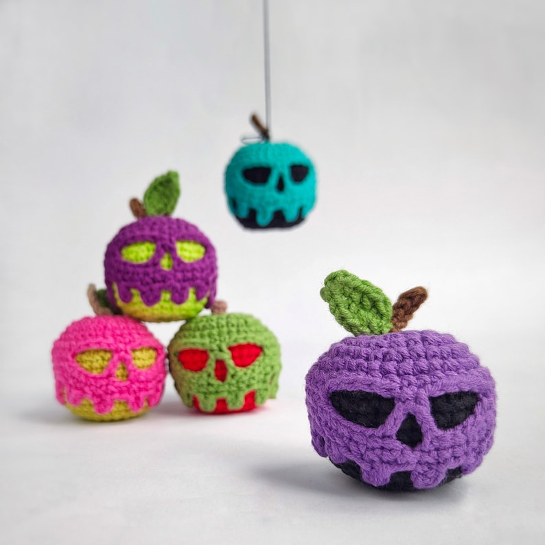 May include: Five crocheted apple ornaments with skull faces. The apples are purple, pink, green, and blue. The ornaments are hanging from a string.