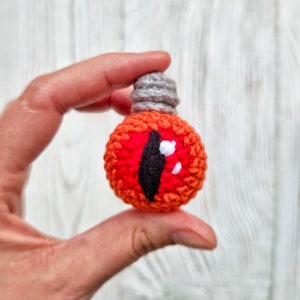 May include: A crocheted lightbulb with a red eye and white pupils. The lightbulb is gray at the top and orange at the bottom.