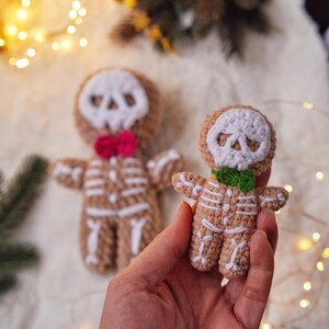 May include: Crocheted skeleton gingerbread men. The figures are tan with white skeleton detailing and skull faces. One has a pink bow tie, the other a green bow tie. The background includes a Christmas tree and fairy lights.